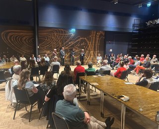 Veel mensen in een zaal die naar een paar sprekers kijken